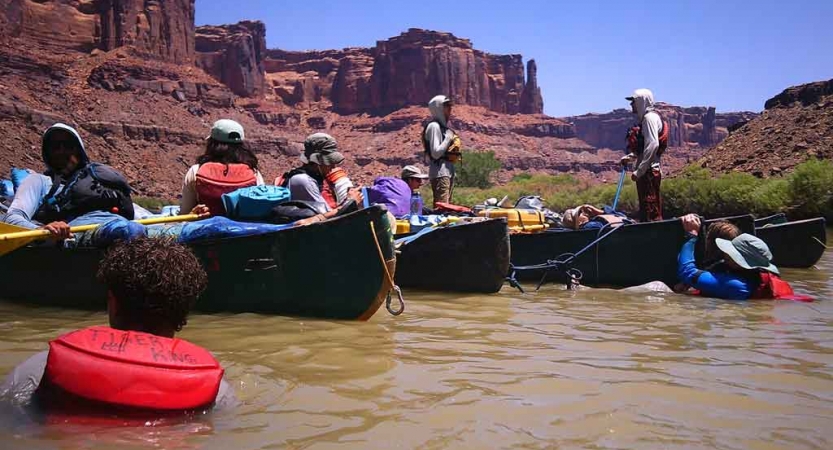 A group of canoes are tethered together while floating in calm water. There are a couple of people in the water. In the background, there are tall, red, canyon walls. 